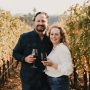 Tyler and Kelly Mullman pose in a vineyard with glasses of wine.